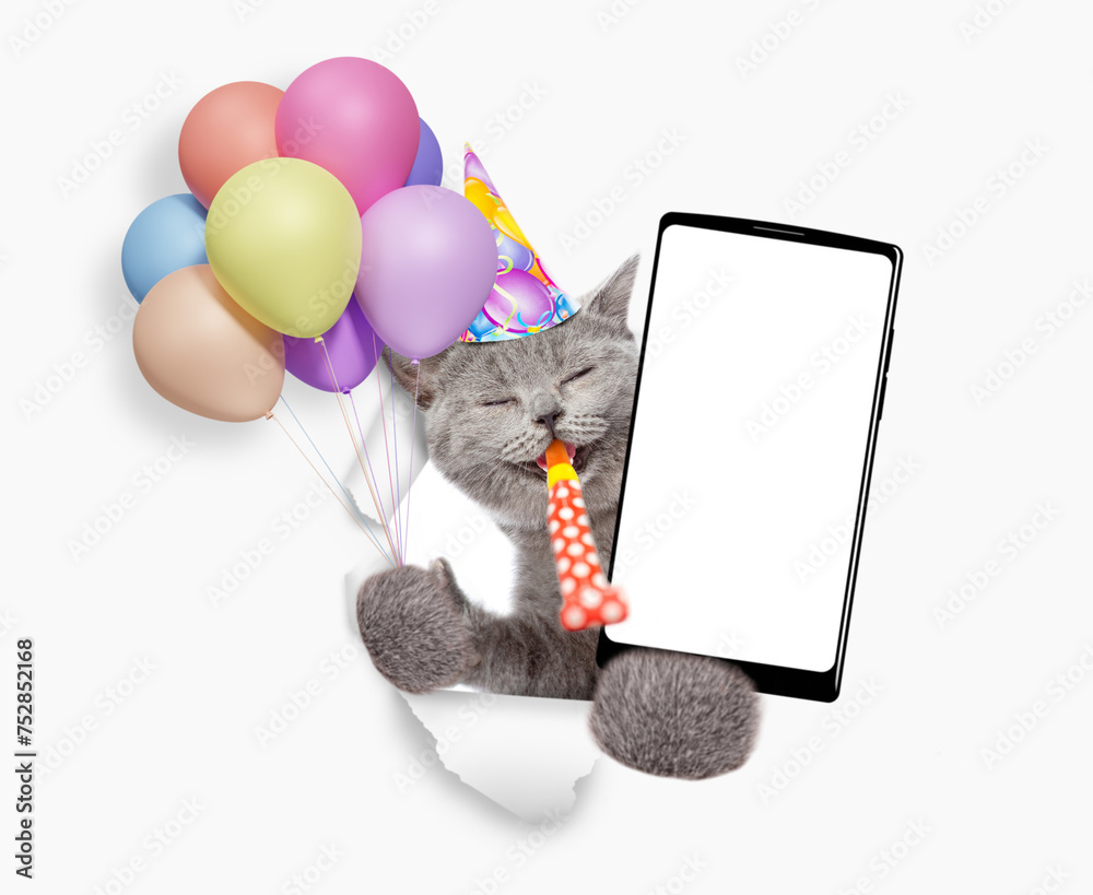 Poster happy kitten wearing party cap holds bunch of balloons, blows in party horn and shows smartphone wit