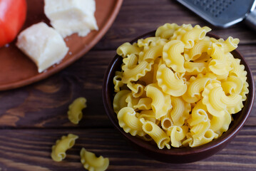 Raw Creste Di Gallo pasta in a ceramic bowl with fresh tomatoes and parmesan cheese on a brown wooden board. Rooster comb. The concept of traditional Italian cuisine. Horizontal orientation.