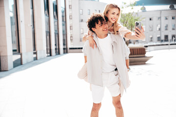 Young smiling beautiful woman and her handsome boyfriend in casual summer clothes. Happy cheerful family. Couple posing in street at sunny day. Female sits at his back. piggyback riding. Take selfie