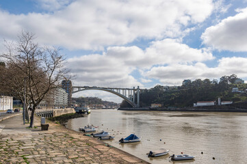 A view East, towards Porto, up the river Douro.  A large, concrete bridge can be seen, along with small boats moored by the river side.  It is sunny and cloudy.