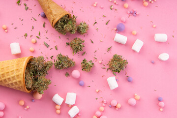 Dry buds of medical marijuana lie on waffle ice cream cones on a pink background.  There are candies and marshmallows . around.  Alternative medical cannabis treatment