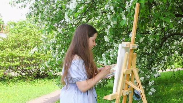 Female artist painting outdoor under the tree in spring blooming park, woman with easel on plein air in floral garden, young girl in summer dress drawing, view from the side