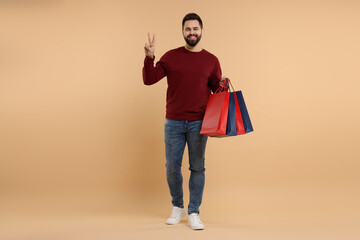 Smiling man with many paper shopping bags showing peace sign on beige background