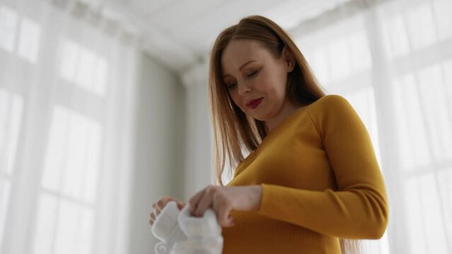 Pregnant woman enjoying expecting baby. Happy female future mother holding child knitted soft white booties in hands with smile, tenderly stroking tummy. Healthy mom pregnancy, expecting baby concept.