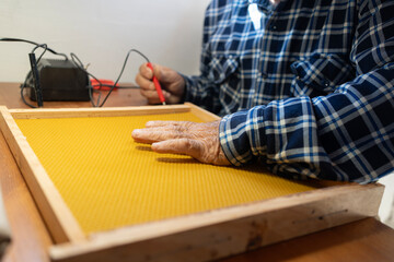 Detail of an old beekeeper's weathered hands gluing sheets of wax on a frame with a small...