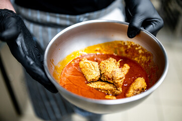 person wearing black gloves, holding a white bowl containing crispy fried chicken wings coated in an orange sauce. vibrant orange sauce clings to the wings, suggesting a flavorful and appetizing dish.