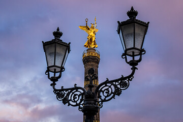 Fototapeta na wymiar Berlin Victory Column, Siegessäule, stands resplendent in the warm glow of the sun, its golden hue illuminated by the evening light, while lanterns frame this iconic monument, adding to its beauty.