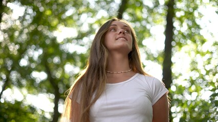 Naklejka na ściany i meble beautiful girl smiling. inner beauty and love for nature concept. beautiful woman stands in a park outdoors in lifestyle a white T-shirt and smiles, face close-up, sun glare on the background