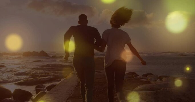 Animation of light spots over biracial couple running at beach