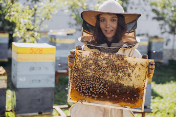 Happy smiling female Beekeeper in protective suit holding honeybee frame with bees at apiary