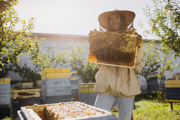 Happy smiling female Beekeeper in protective suit holding honeybee frame with bees at apiary