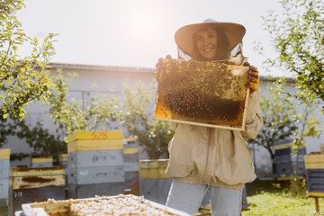 Happy smiling female Beekeeper in protective suit holding honeybee frame with bees at apiary
