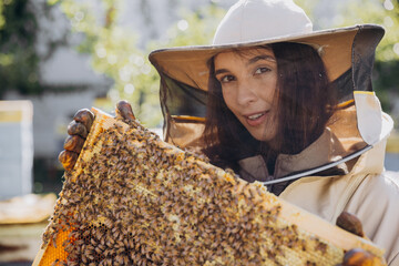 Happy smiling female Beekeeper in protective suit holding honeybee frame with bees at apiary