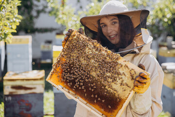 Happy smiling female Beekeeper in protective suit holding honeybee frame with bees at apiary