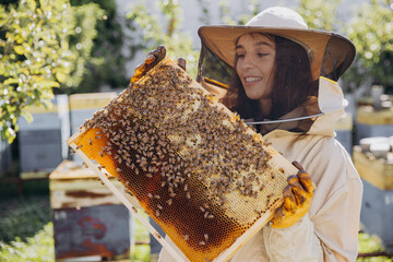 Happy smiling female Beekeeper in protective suit holding honeybee frame with bees at apiary