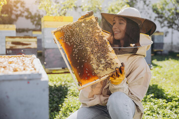 Happy smiling female Beekeeper in protective suit holding honeybee frame with bees at apiary