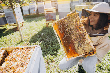 Happy smiling female Beekeeper in protective suit holding honeybee frame with bees at apiary