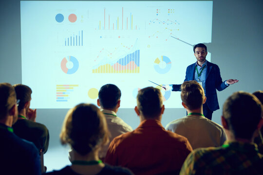 Businessman, speaker delivering presentation to attentive crowd with projection of colorful statistical graphs behind. Concept of business, startup, leadership and personal development courses.