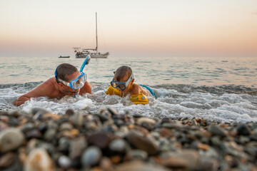 Young man with his son 4 years old swim near shore in masks with snorkels sunset, background yacht.