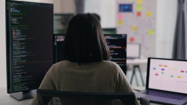 A female developer works attentively with code on multiple monitors in a well-organized office space, surrounded by digital workflow charts.