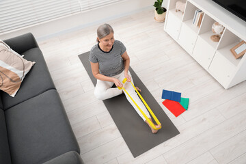 Senior woman doing exercise with fitness elastic band on mat at home, above view