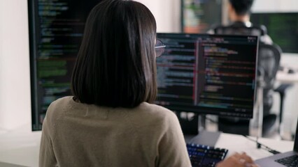 Rear view of a female programmer intensely coding on dual monitors in a dynamic office environment, possibly engaged in cybersecurity or software development.