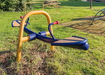 An empty colourful seesaw for kids stands in an outdoor playground with grass.