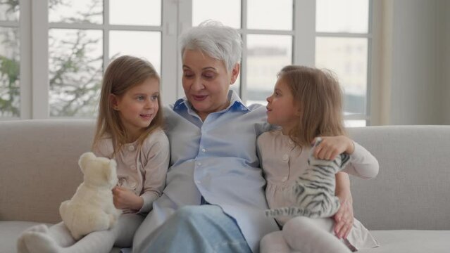 Grandmother with granddaughters resting. Happy relaxed older adult granny embracing preschool children two twins granddaughters cuddling talking rest on couch at home. Family love, generation concept.