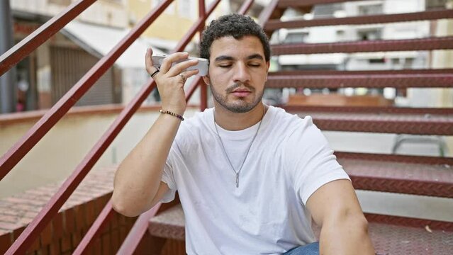A young mixed-race man with a beard using a smartphone on urban outdoor stairs, displaying casual style.