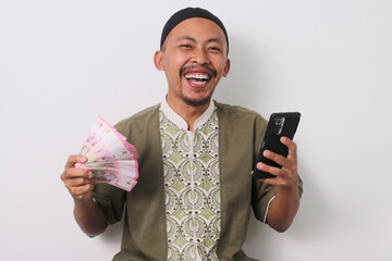 Excited Indonesian Muslim man in koko shirt and peci holds a phone and Indonesian rupiah banknotes,...