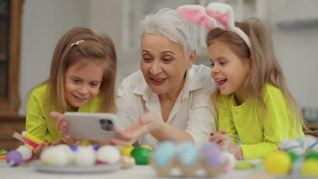Easter grandmothers with granddaughters. Smiling grandmother with twins grandchildren watching cartoons on phone in rabbit bunny ears, talking, celebrating together close up. Easter holiday concept.