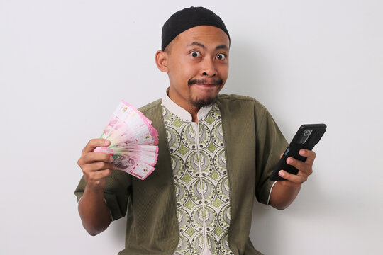 Excited Indonesian Muslim man in koko shirt and peci holds a phone and Indonesian rupiah banknotes, celebrating financial success. Isolated on white background