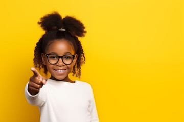 Black little girl 10 years old in a white T-shirt and glasses on a yellow background schoolgirl with her finger to the side - obrazy, fototapety, plakaty