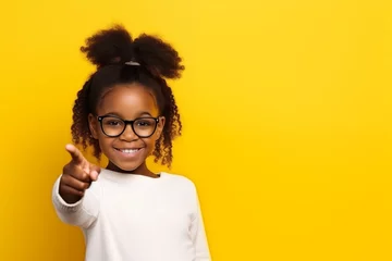 Foto op Aluminium Black little girl 10 years old in a white T-shirt and glasses on a yellow background schoolgirl with her finger to the side © Nelly