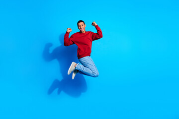 Obraz premium Full size photo of energetic delighted man jump raise fists empty space ad isolated on blue color background