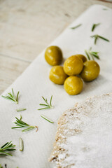 Close up olives on top of the white wooden table next to the raw dough with rosemary leaves.