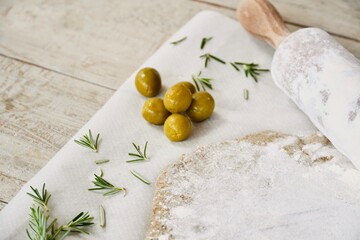 Close up olives on top of the white wooden table next to the raw dough with rosemary leaves.