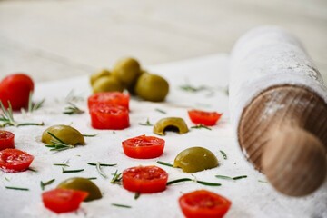 Close up olives and cherry tomatoes on the white wooden table next to the raw dough with rosemary...