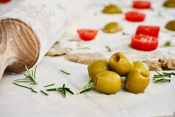 Close up olives on the white wooden table next to the raw dough with cherry tomatoes and rosemary leaves.