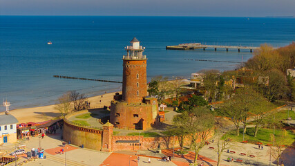 A sunny day in February at Kołobrzeg port, Poland. The image captures a red brick lighthouse, tourists strolling on the sandy beach, and a distant pier, with calm sea waters. - obrazy, fototapety, plakaty