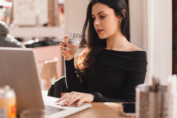 Smart caucasian woman with glass of water in hand studying with laptop. Student teen girl sitting in cafe drinks glass of water for body hydration while studying. Healthy lifestyle theme.