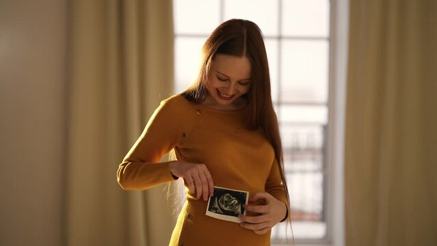 Pregnant woman looking ultrasound report. Happy pregnant female watching her ultrasound report and touching her abdomen, admiring sonography picture of her unborn baby. Expecting baby lifestyle.