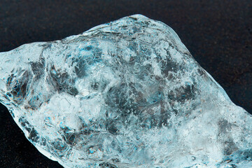 Close-up of a clear, textured ice chunk that looks like a diamond on a dark surface, highlighting...