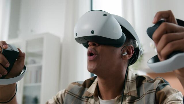 Excited African American man ethnic guy gamer video game addict male at home playing vr gaming play online using modern virtual reality meta world simulation wearing 3d helmet glasses hold controllers