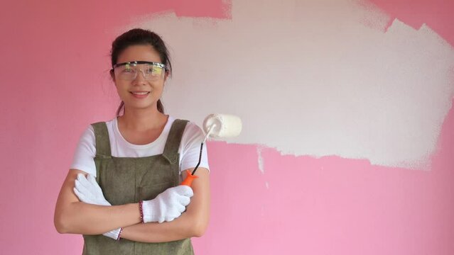 a smiling Asian woman painting the wall of her house with a roller brush. Asian woman changing the wall color of her house to white home interior ideas	