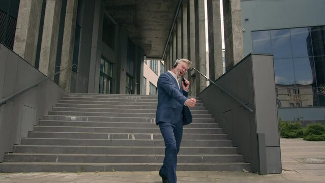 Happy senior man in headphones with phone mp3 music app walking down stairs near office business company building moving dancing old businessman middle-aged Caucasian mature employer listen song fun