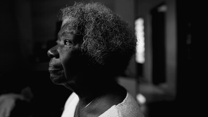 One introspective black senior woman in solitude at home in black and white. Monochromatic close-up face of an African American lady staring at window with thoughtful emotion