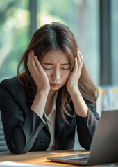 Exhausted young Asian office worker, woman in formal clothes sitting at desk, looking tired, eyes closed, hands on her head. Generative AI