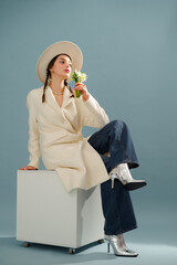 Fashionable confident woman wearing white hat, elegant boucle coat, wide leg jeans, silver boots, holding spring bouquet of snowdrops, posing on blue background. Full-length studio portrait
- 753061766