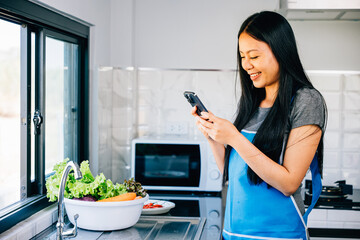 A smiling Asian woman cooks vegetables in a wooden kitchen checking her smartphone for a cooking tutorial. Cheerfully browsing fruits she blends technology into her culinary journey. mobile phone